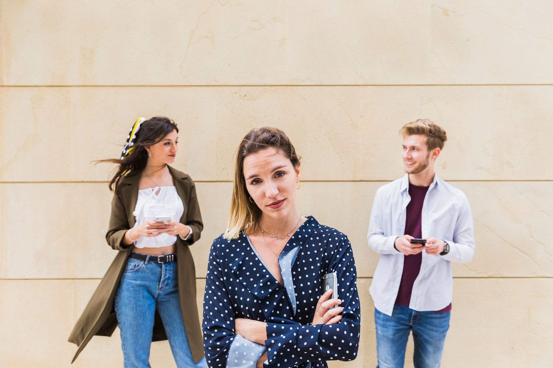 A woman is standing in front of a wall while two men look at their phones.