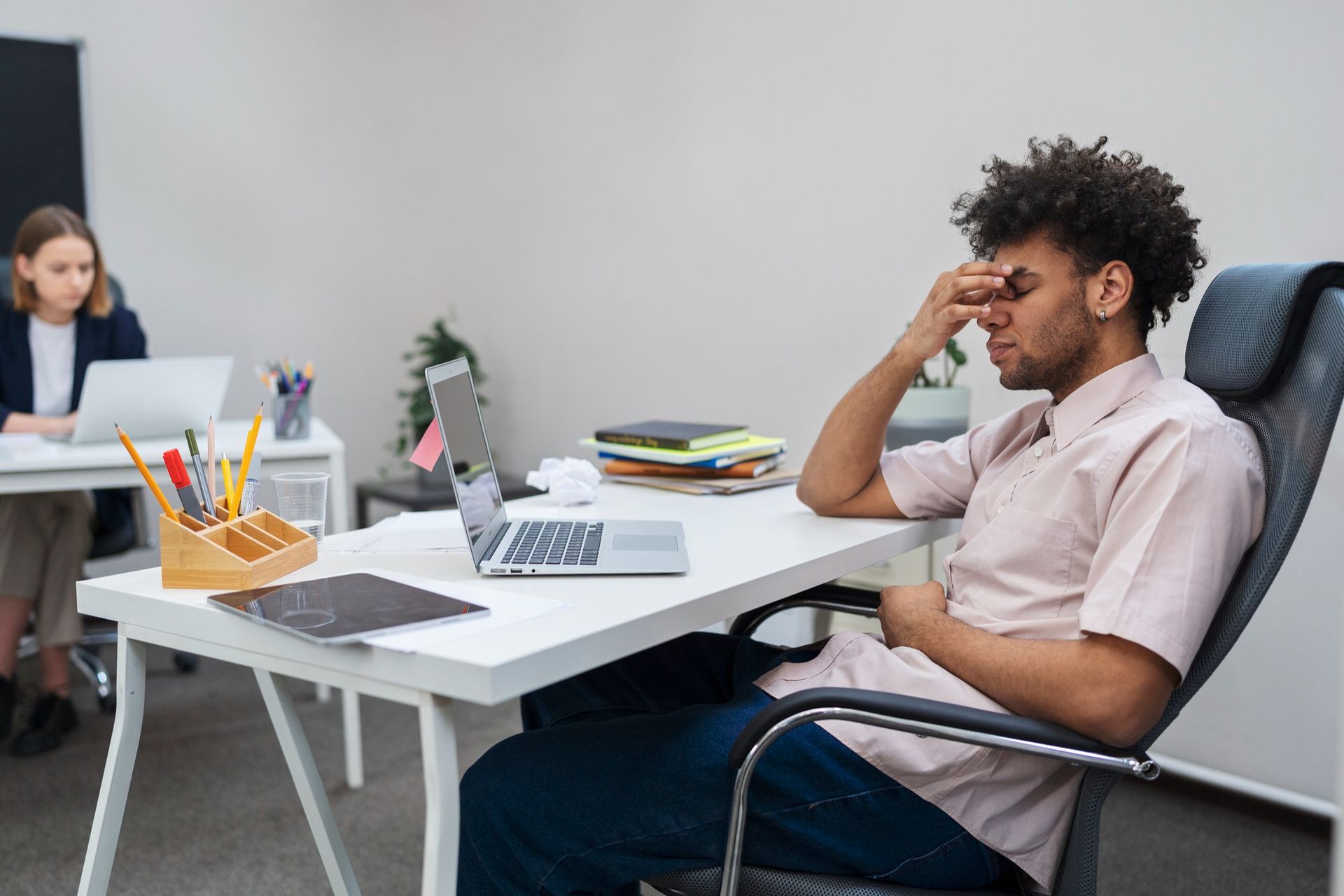 Man at desk with hand on face, appears stressed, laptop open. Another person works at a desk behind.