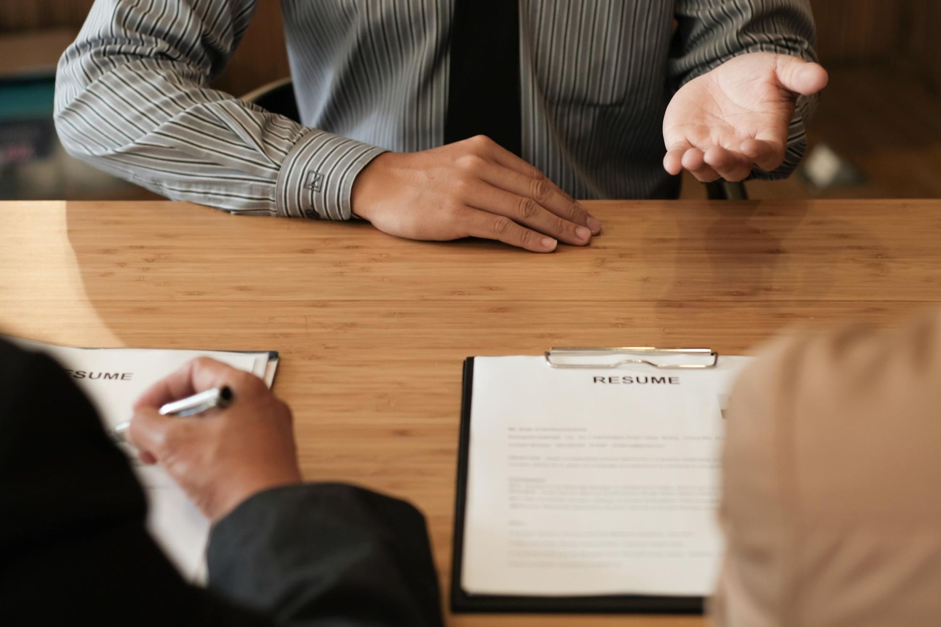 A person gesturing while seated at a wooden table across from two people, resumes in view.