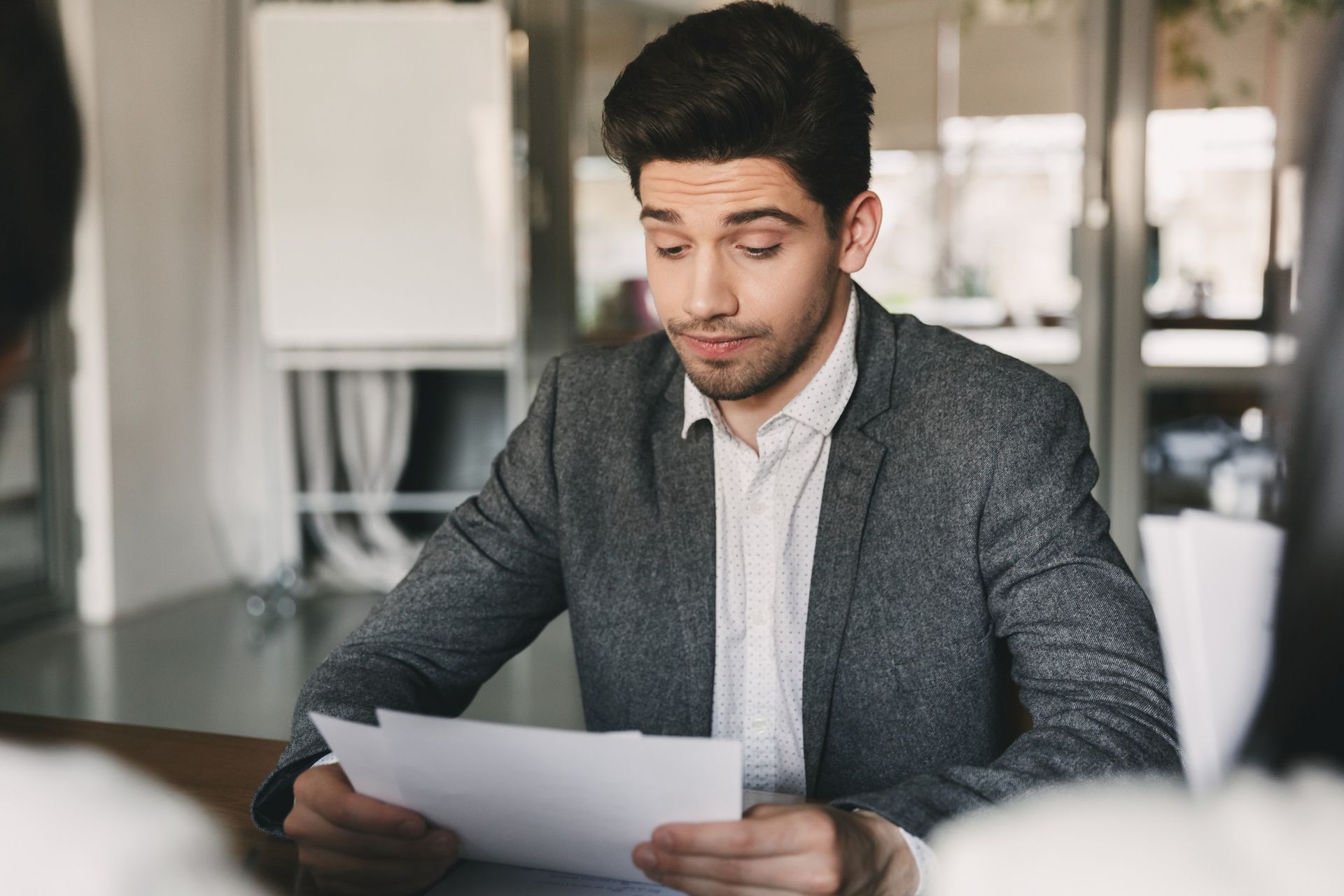 Man in gray blazer reads papers at a table in an office.