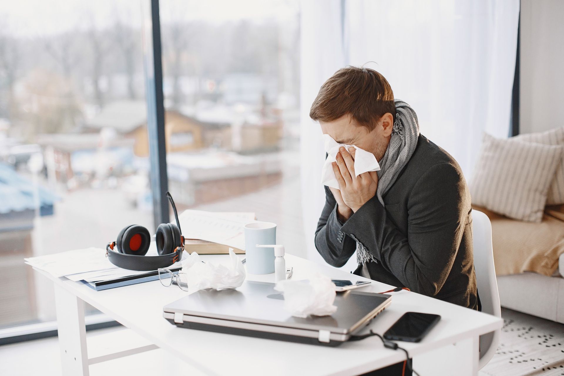 Man seated at a desk, blowing his nose with a tissue; laptop, headphones, and used tissues are visible.