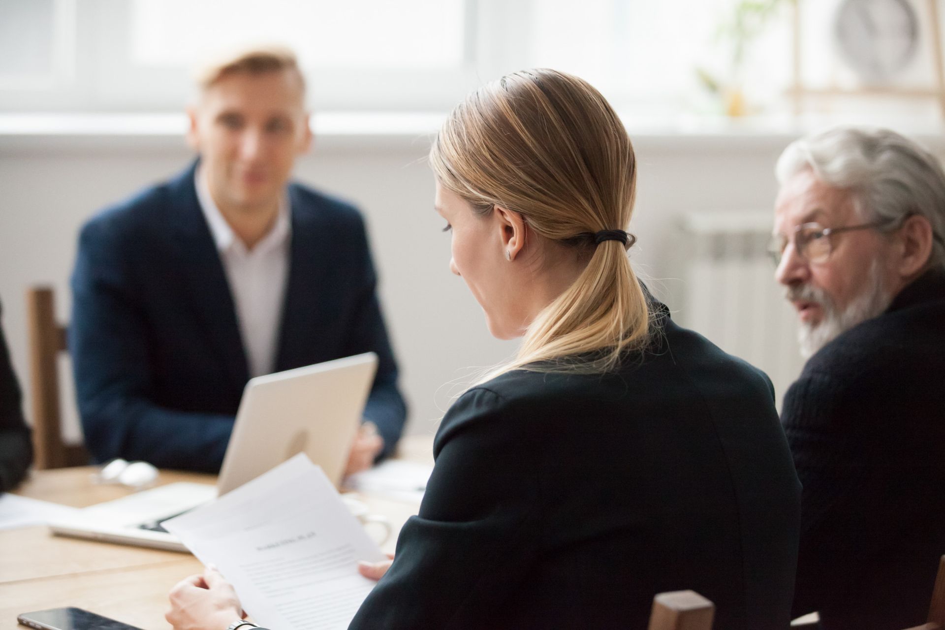 Business meeting: Woman reads document, two men sit at the table, one with a laptop.