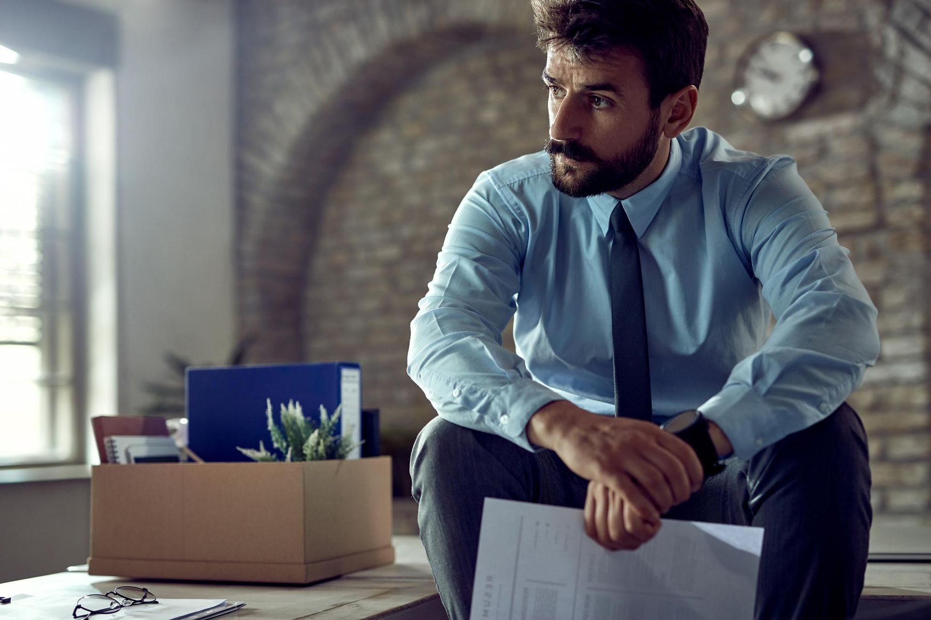 A dejected person in professional attire sits with a box of personal belongings, signifying a job loss.