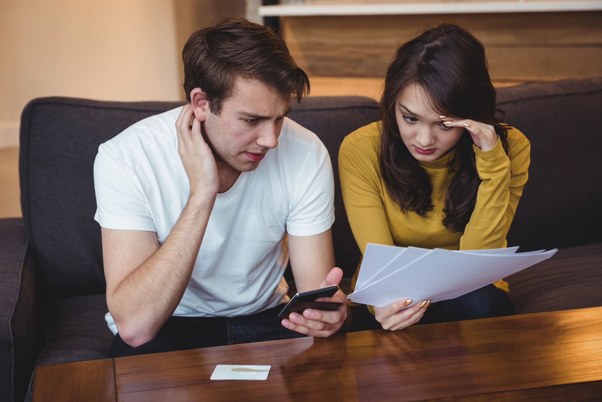 A man and a woman are sitting on a couch looking at papers and a cell phone.