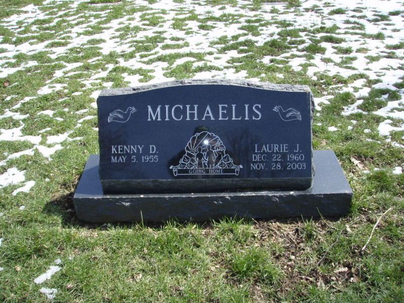Black headstone for Kenny D. Michaelis and Laurie J. Michaelis in a grassy field with a few patches of snow.