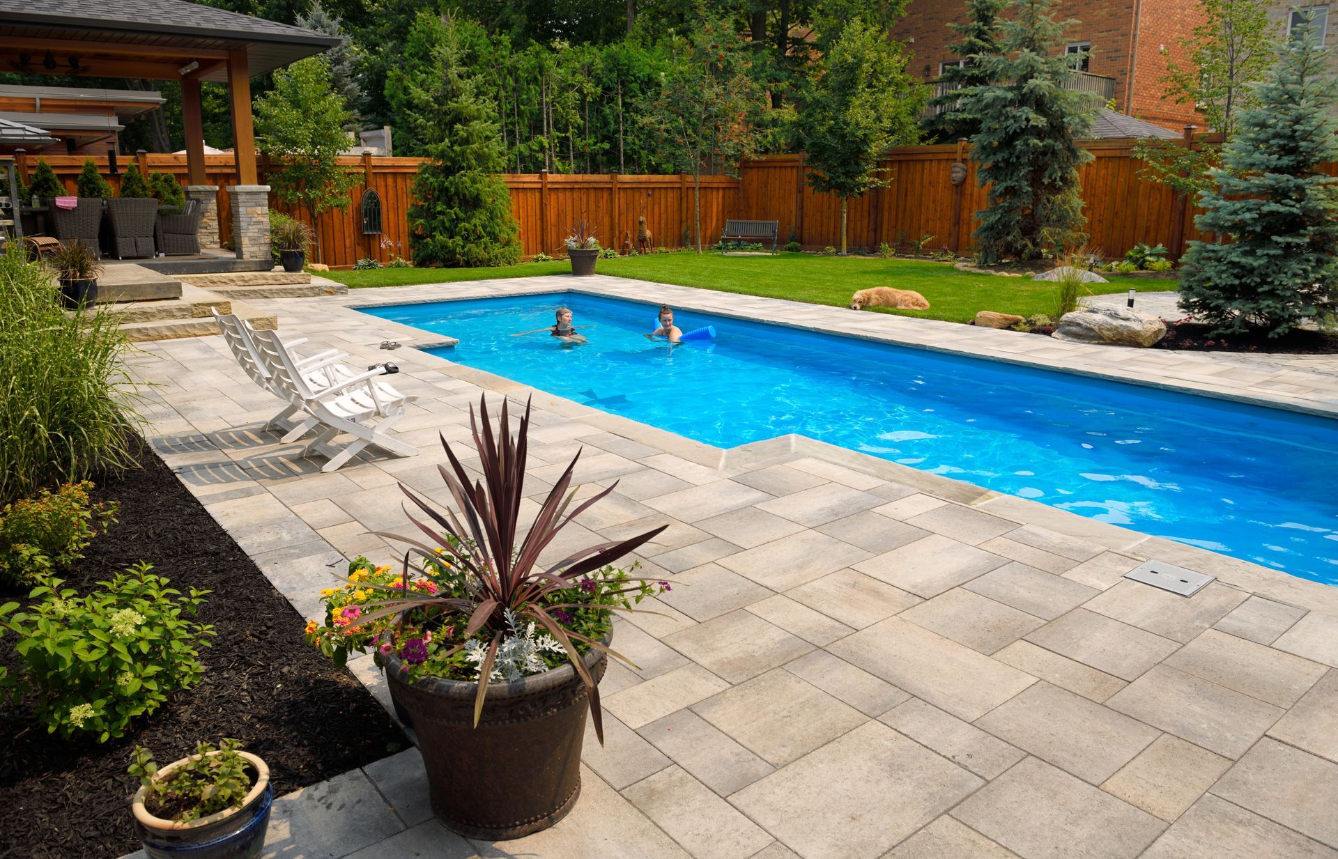 Backyard swimming pool with paved patio, lounge chairs, and people relaxing in the water.