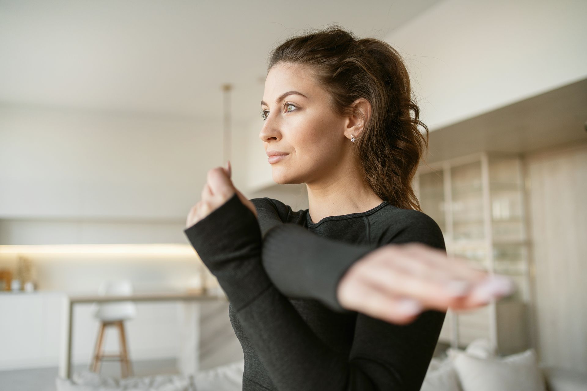 Woman in black workout gear stretching indoors, looking away.