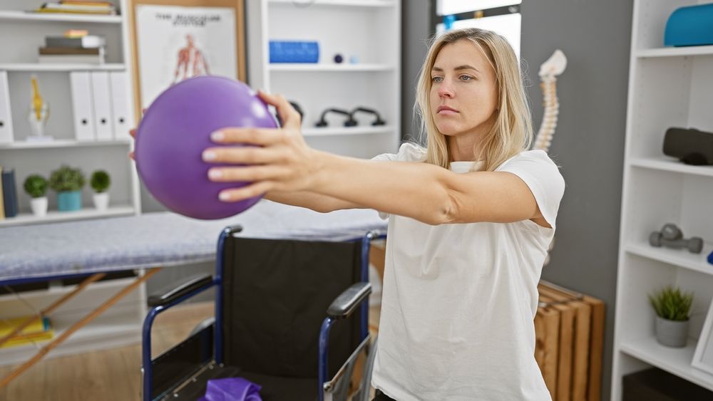 Woman in a bright room holding a purple exercise ball with arms extended.