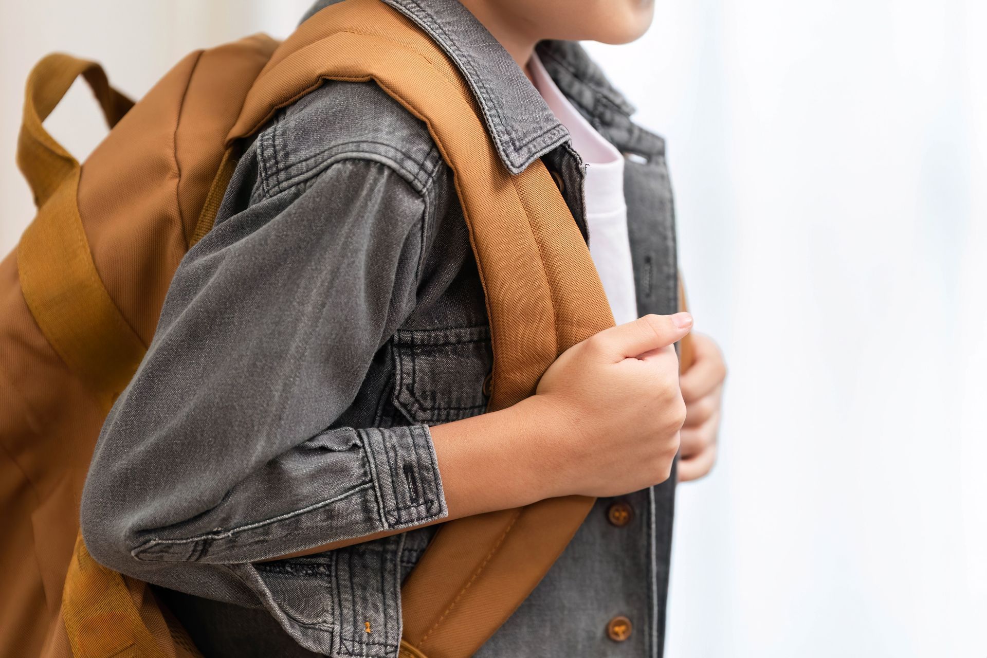 Child in denim jacket with a brown backpack, holding the straps, indoors.