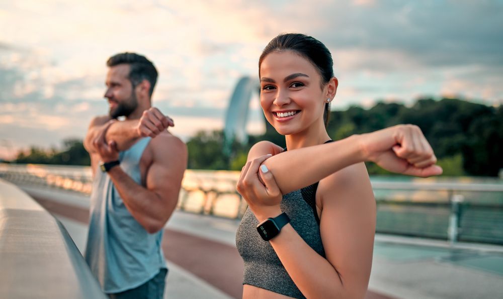 Two people stretching on a bridge at sunset, smiling in workout clothes.