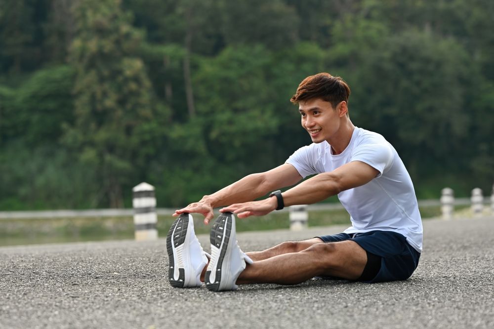 Man sitting on a road stretching toward his toes outdoors, wearing a white shirt and black shorts.