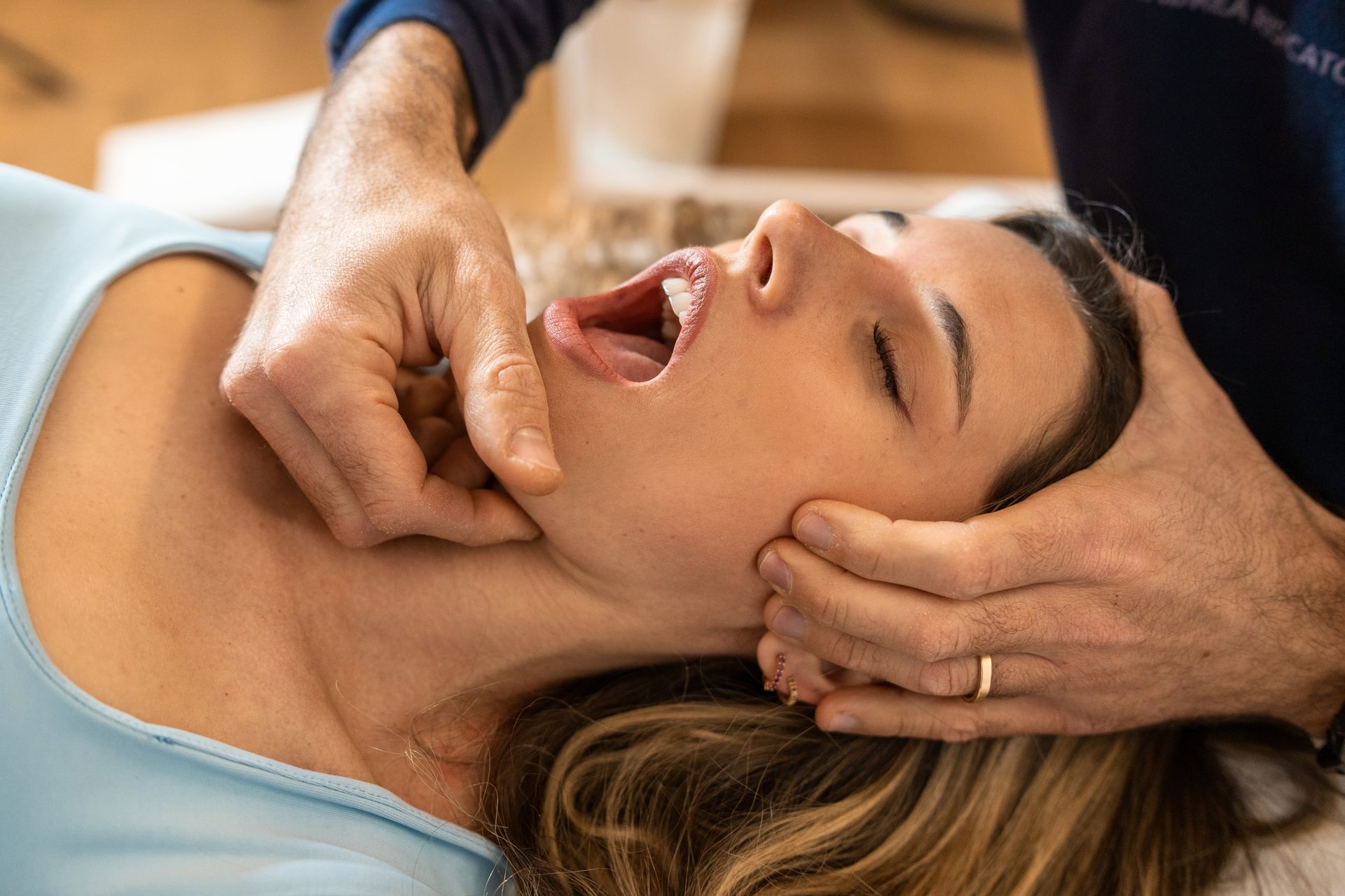 Woman receiving jaw therapy. Man's hands manipulate her jaw and neck. Her mouth is open.