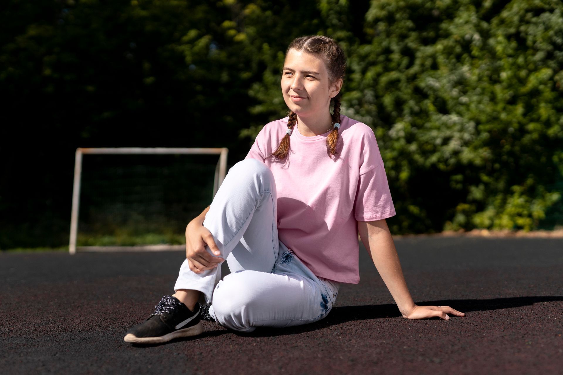 Woman with braids in pink shirt and jeans, sitting on a dark surface, looking off to the side.