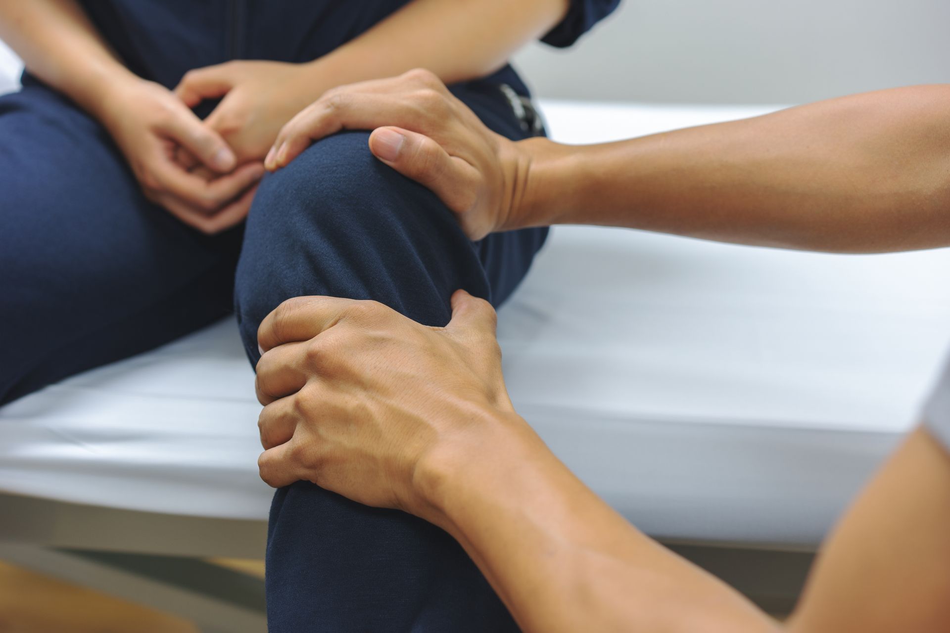 A person is sitting on a bed getting their knee examined by a doctor.