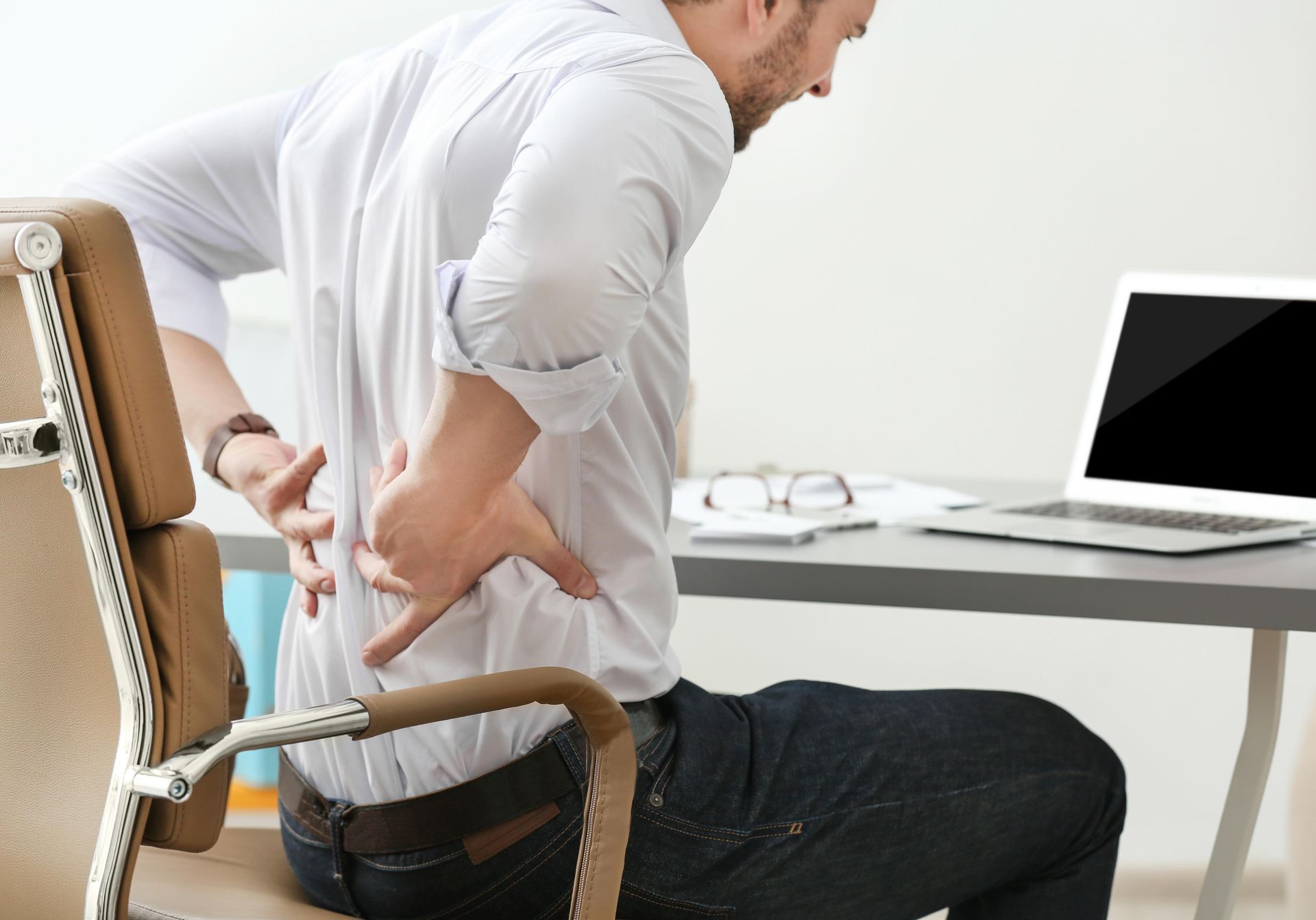 A man is sitting in an office chair holding his back in pain.