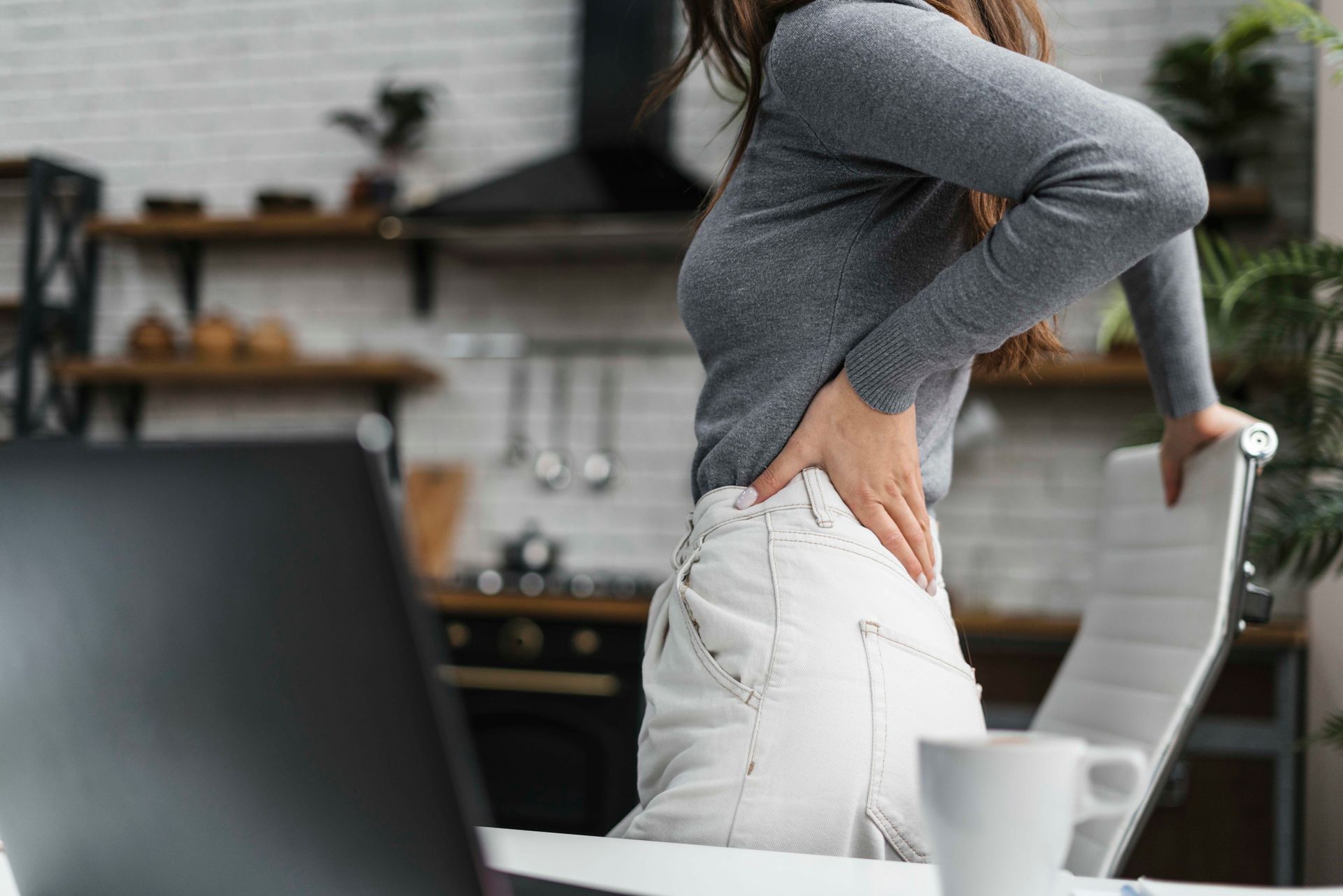 A woman is sitting at a desk with a laptop and holding her back in pain.