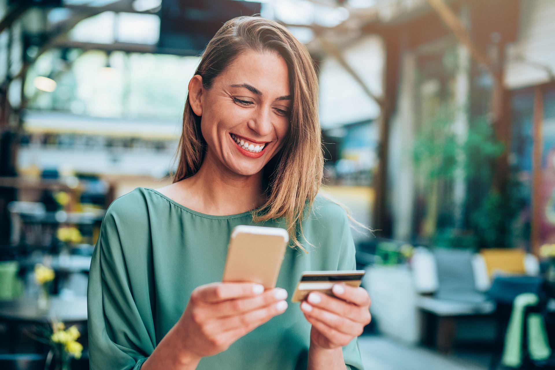 A woman is smiling while holding a credit card and a cell phone