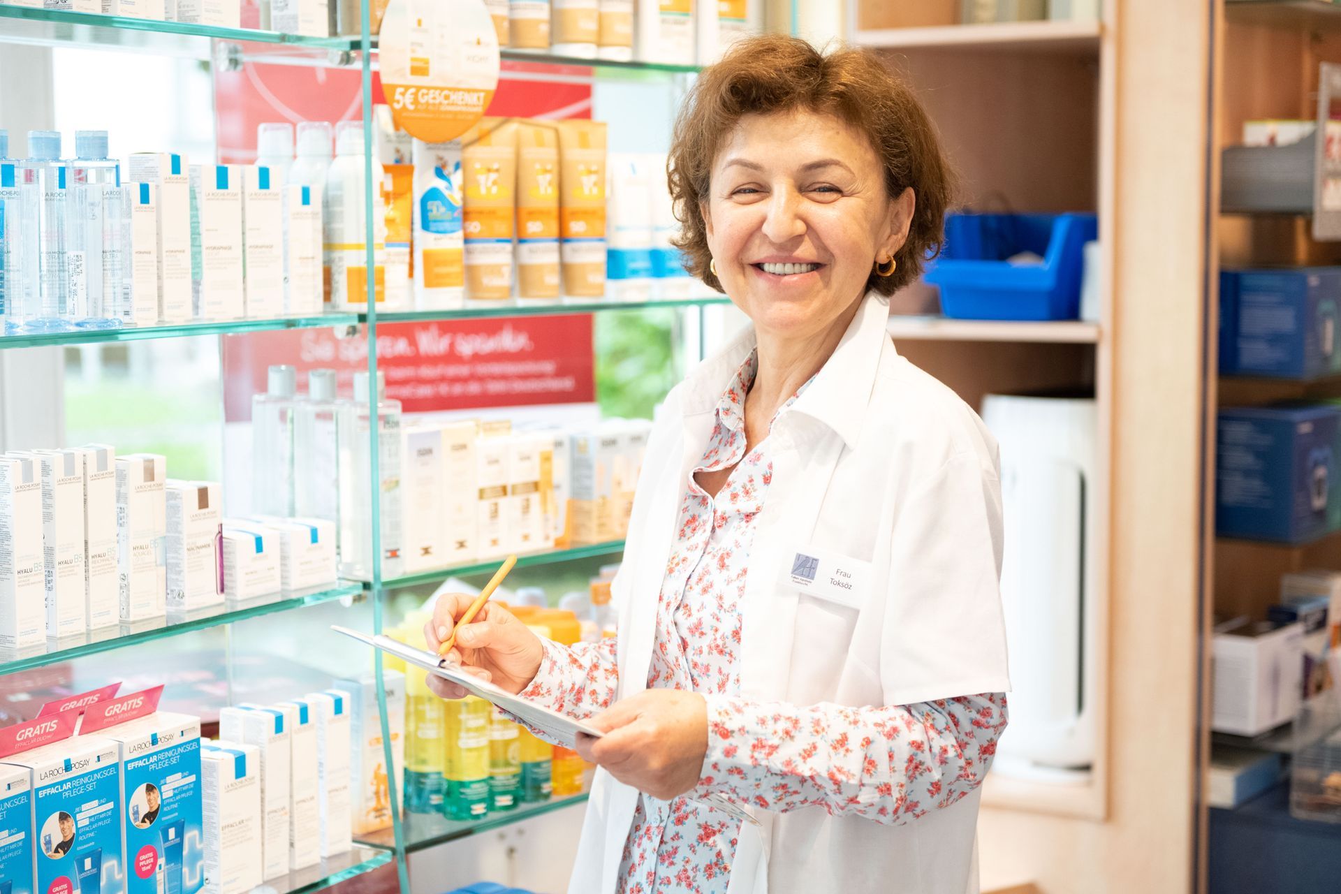 Eine Apothekerin lächelt und hält in einer Apotheke ein Klemmbrett in der Hand.