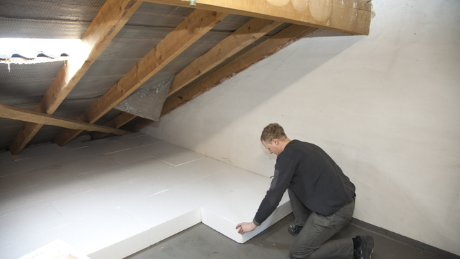 A person in a dark sweater kneels in an unfinished attic, installing white insulation panels onto the floor.