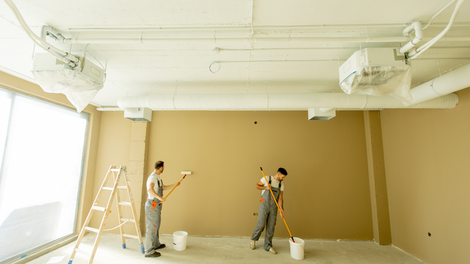 Two people wearing work overalls paint the beige walls of a large room with roller brushes.