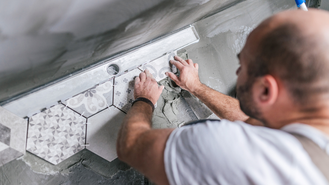 A tiler installs grey and white patterned hexagonal floor tiles with thin-set mortar next to a linear shower drain.
