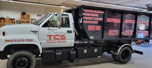 White TCS dumpster truck with red logo, parked indoors. Text includes phone numbers and services offered.