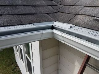 Damaged white gutter and gray roof corner of a house. Metal gutter guards are visible.