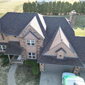 Brown shingled roof of a two-story house with a brick chimney and stone facade; an aerial view.