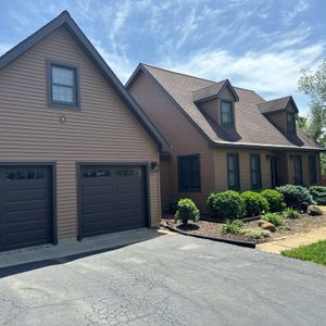 Two-story house with a brown exterior, dark garage doors, and a paved driveway under a blue sky.