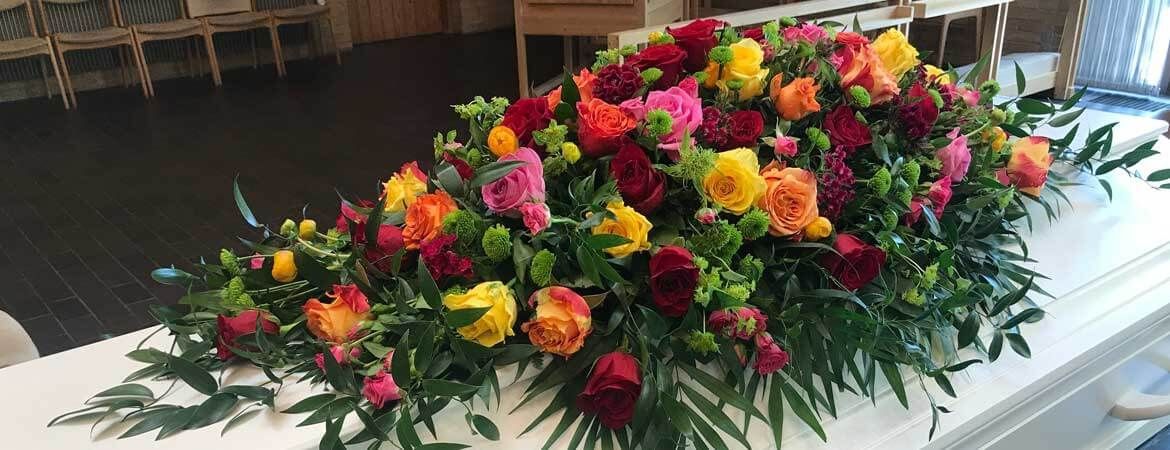 A colorful floral arrangement atop a white coffin, in a room with chairs and light streaming in.