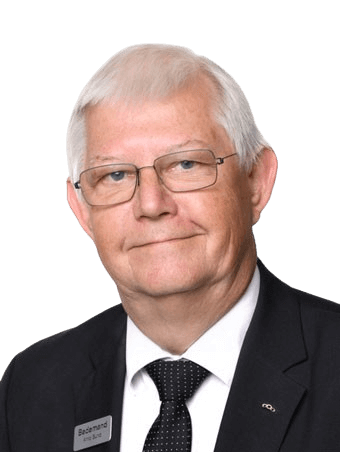 Portrait of an older man with glasses wearing a suit, smiling. Name tag visible.