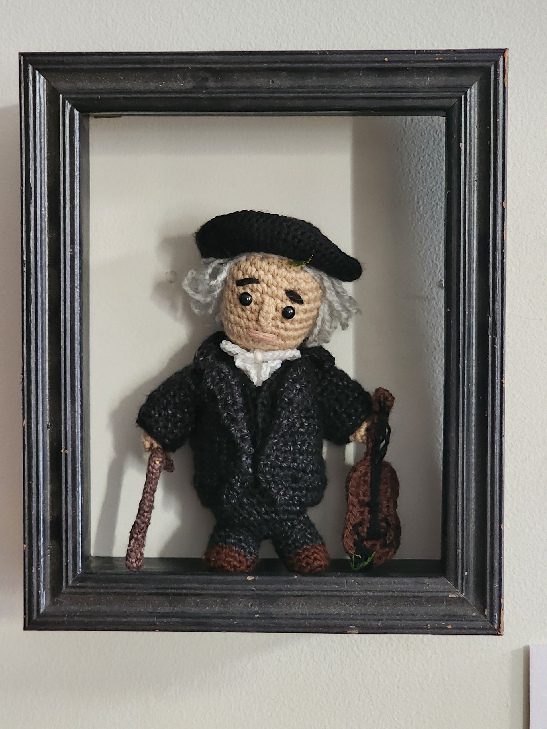 Framed knitted doll in black hat and coat, standing with a cane in a shadow box.