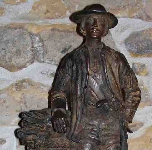 Bronze statue of a man in a hat standing against a stone wall, with a bundle beside him