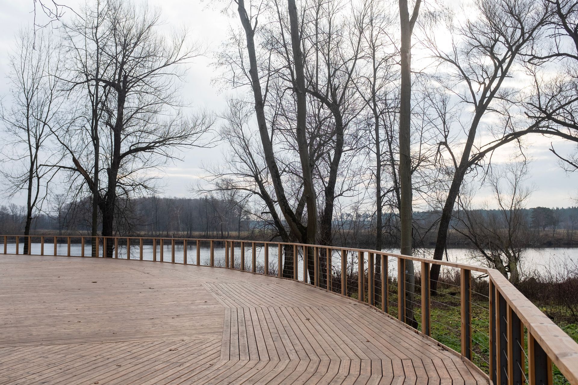 Curved wooden boardwalk beside a lake with bare trees and cloudy sky