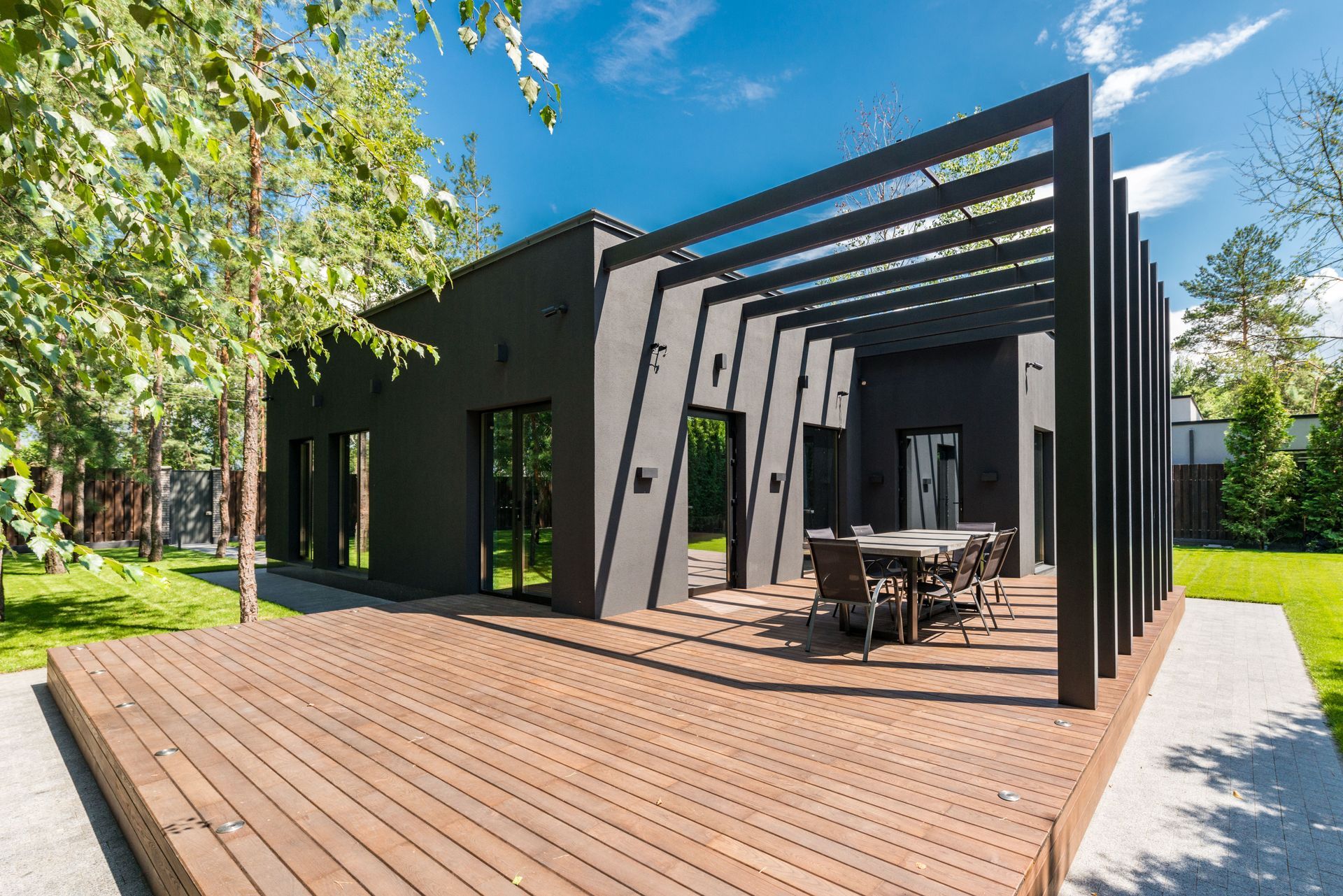 Modern black house with wooden deck, patio seating, and pergola in a sunny yard