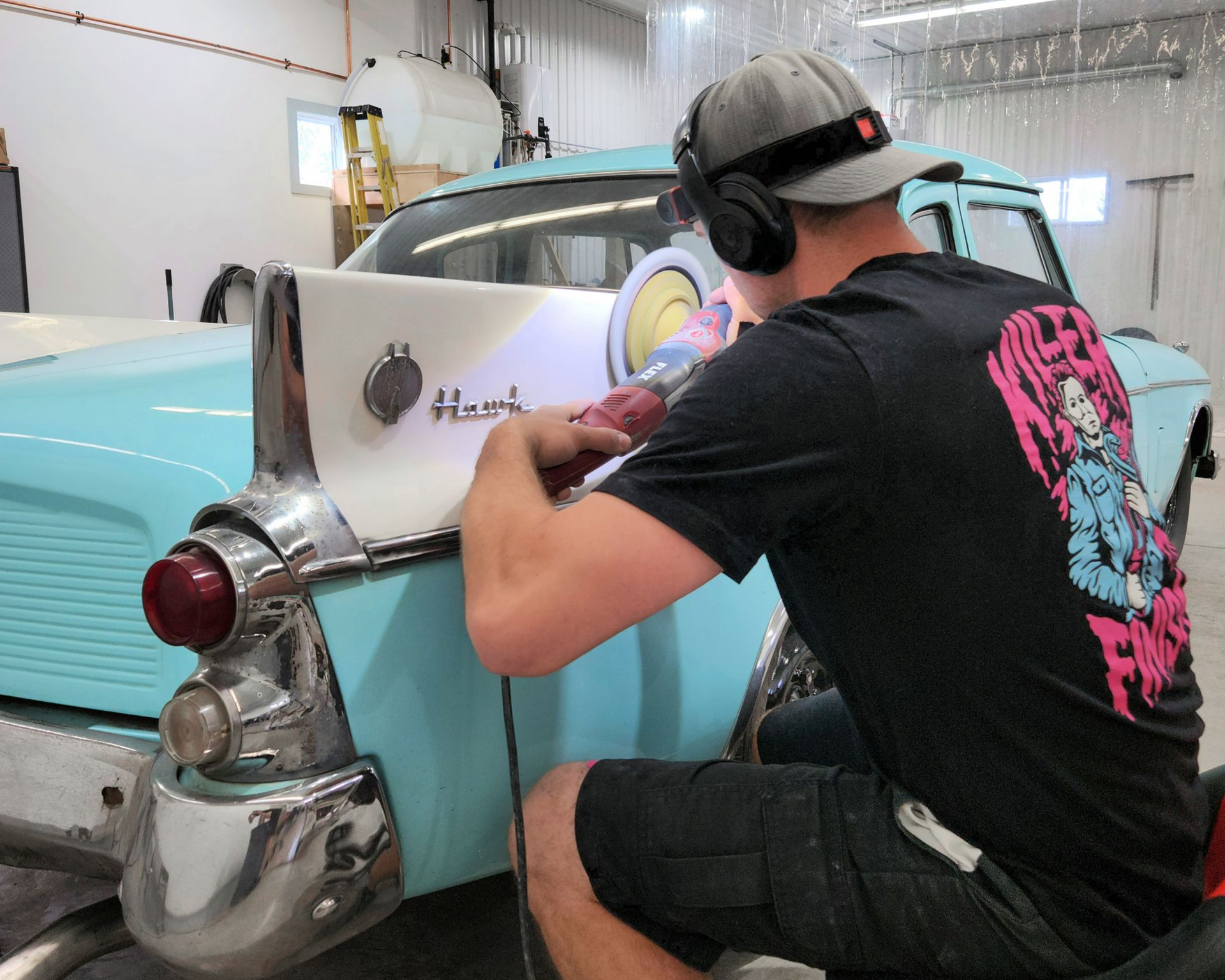 Person welding on a classic turquoise and white car. The man wears headphones and a graphic tee in a shop setting.