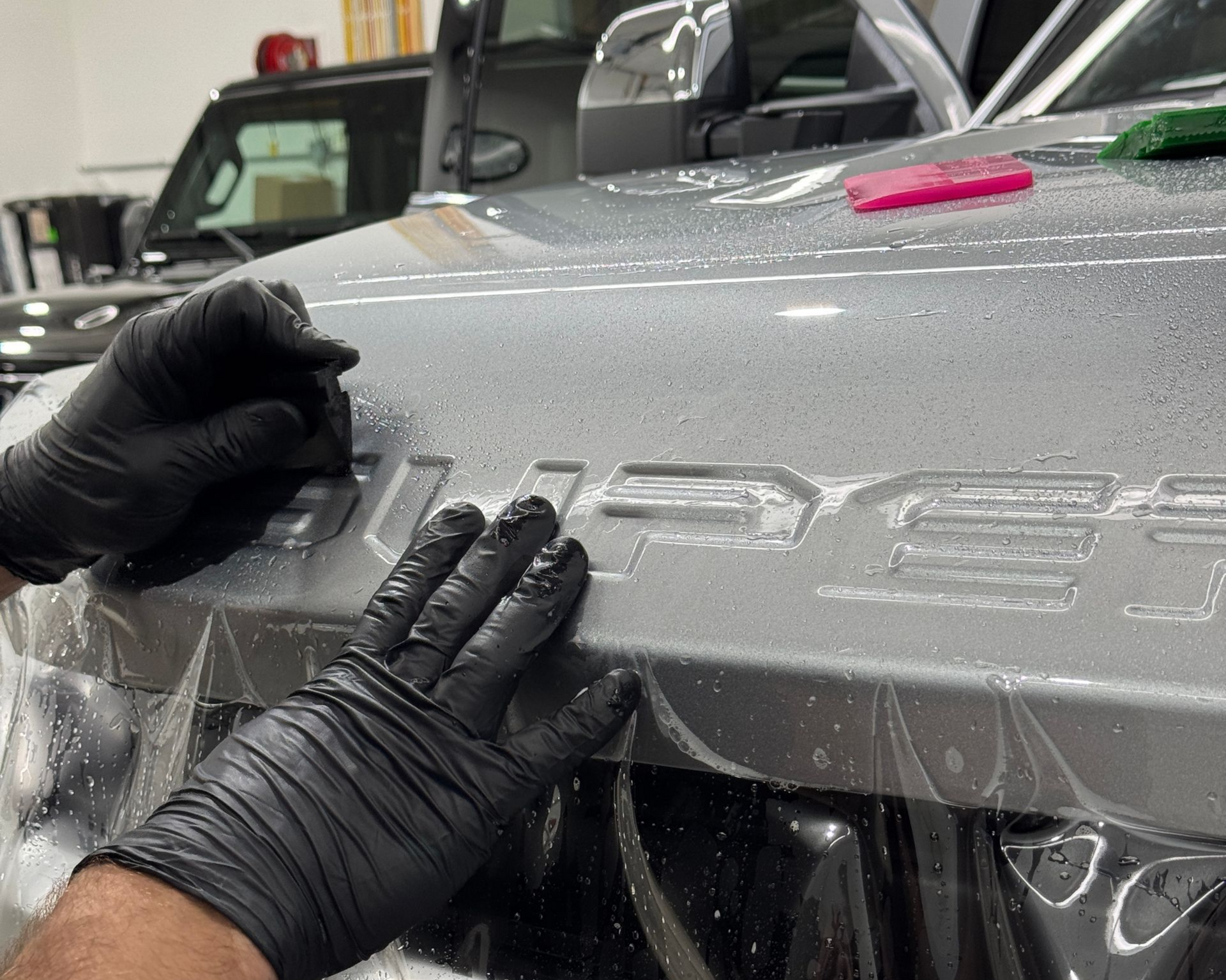 Hands in black gloves applying a clear film to a car hood with raised lettering.