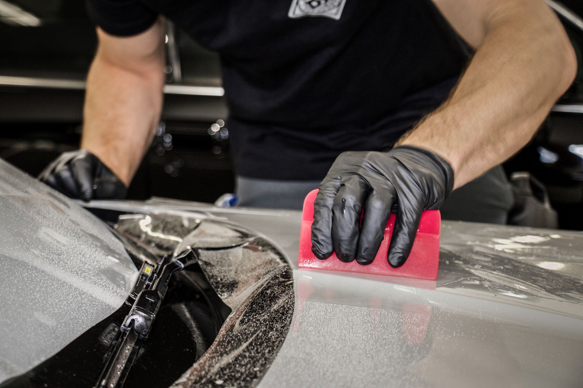 A man wearing black gloves is cleaning a car with a red squeegee.