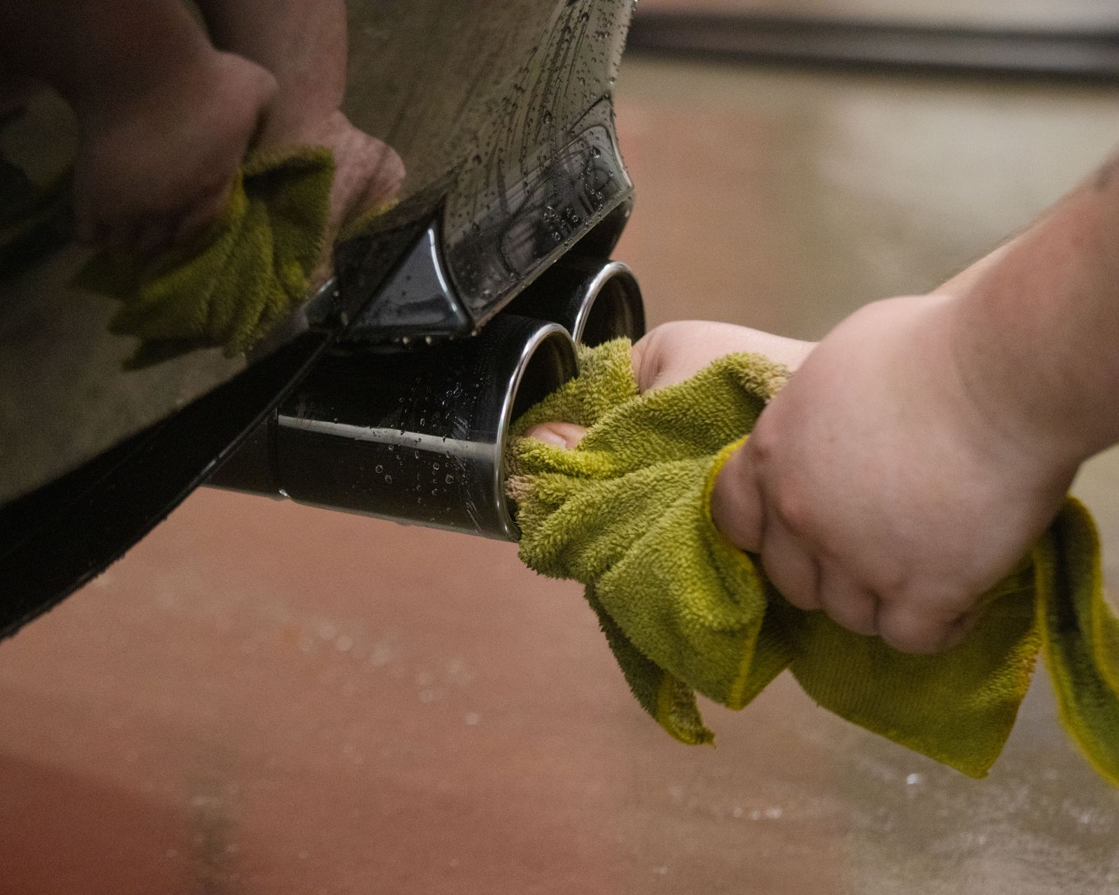 Hands wiping exhaust pipes of a vehicle with a yellow cloth.