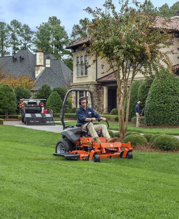 A Man Is Riding a Lawn Mower in Front of A House — CBS Chainsaw & Brushcutter Specialists - HUSQVARNA in Laurieton, NSW