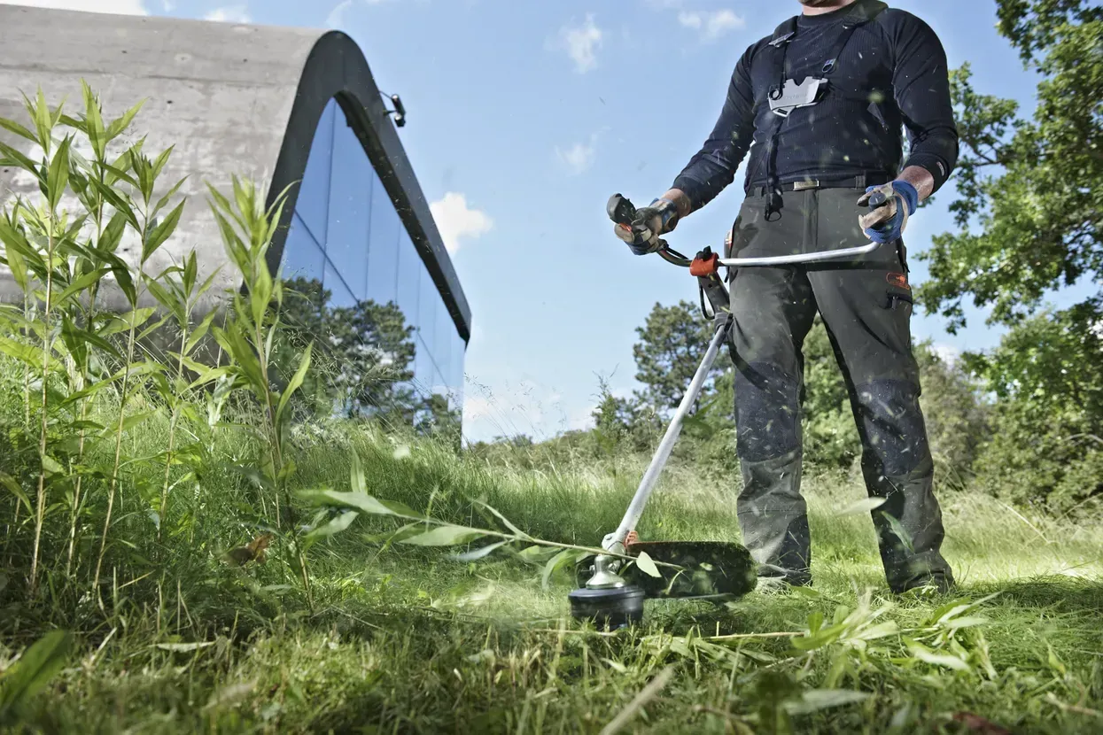A Man Is Using a Grass Trimmer Next To A Building — CBS Chainsaw & Brushcutter Specialists - HUSQVARNA in Wauchope, NSW
