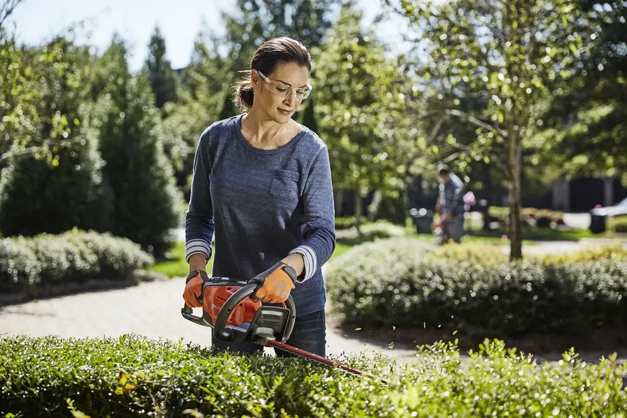 A Woman Is Cutting a Hedge with A Hedge Trimmer — CBS Chainsaw & Brushcutter Specialists - HUSQVARNA in Wauchope, NSW