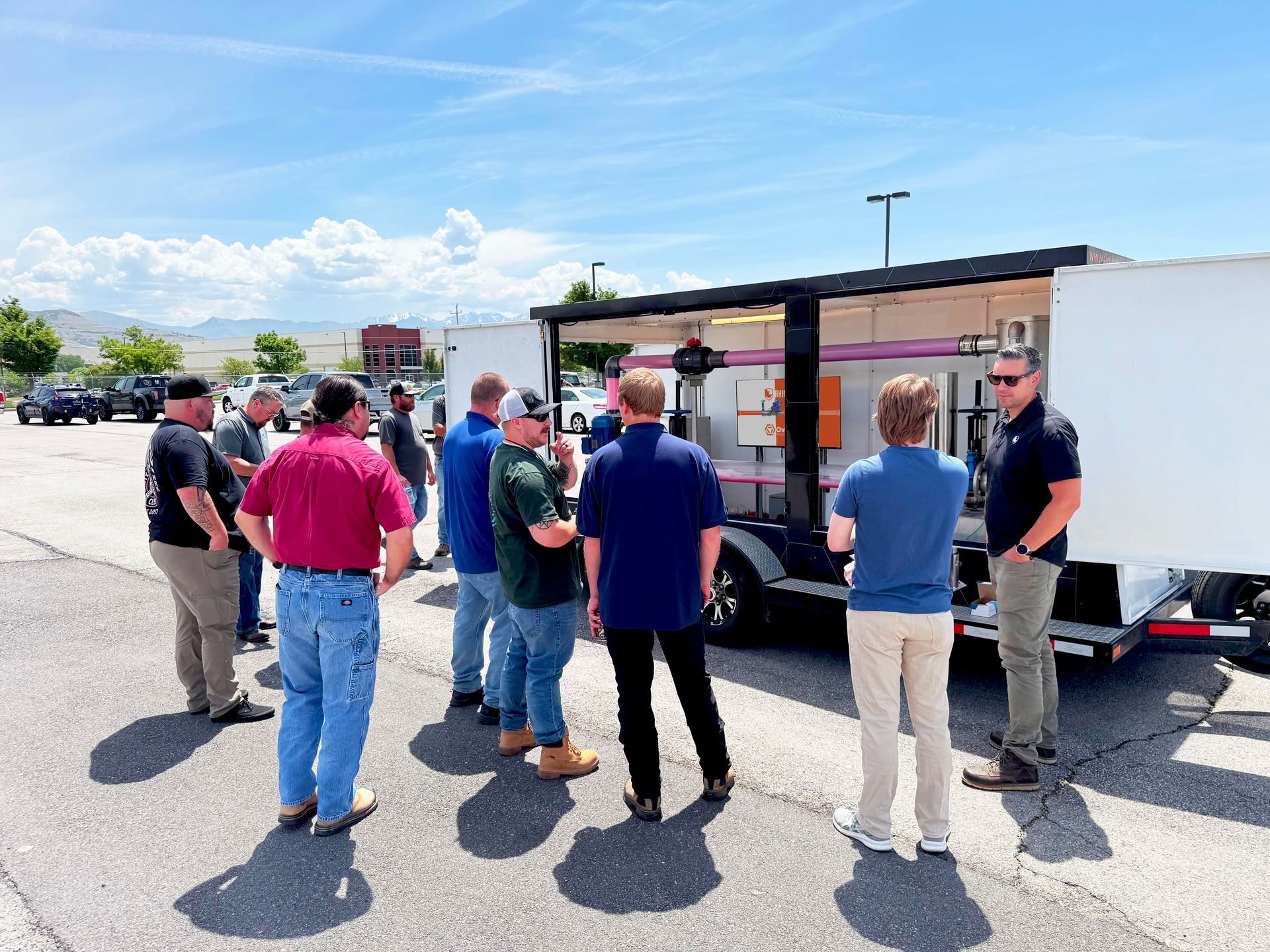 A group of men in casual attire gather around a trailer, likely for a demonstration or training outdoors on a sunny day.
