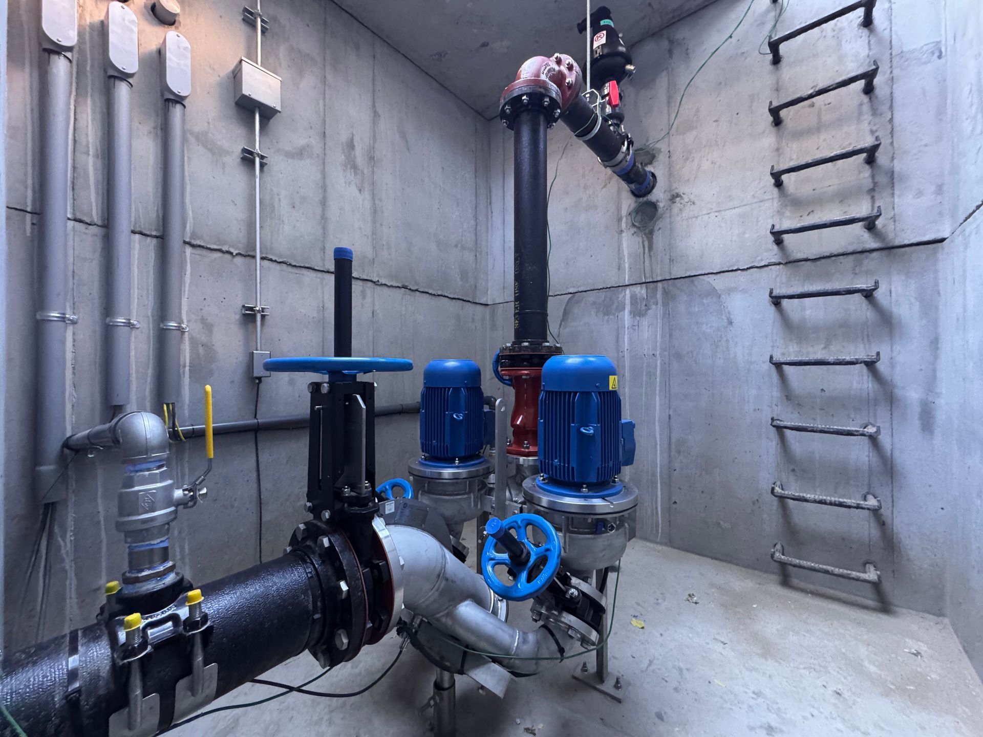 Industrial pump and piping system in a concrete utility chamber with blue motors and a ladder on the wall