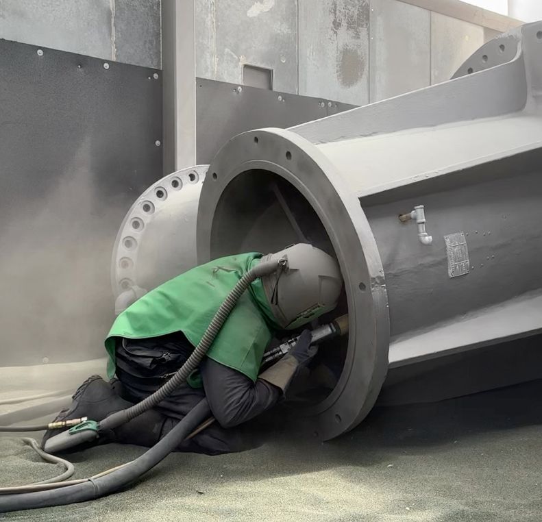 A man wearing a green vest is sandblasting a large pipe