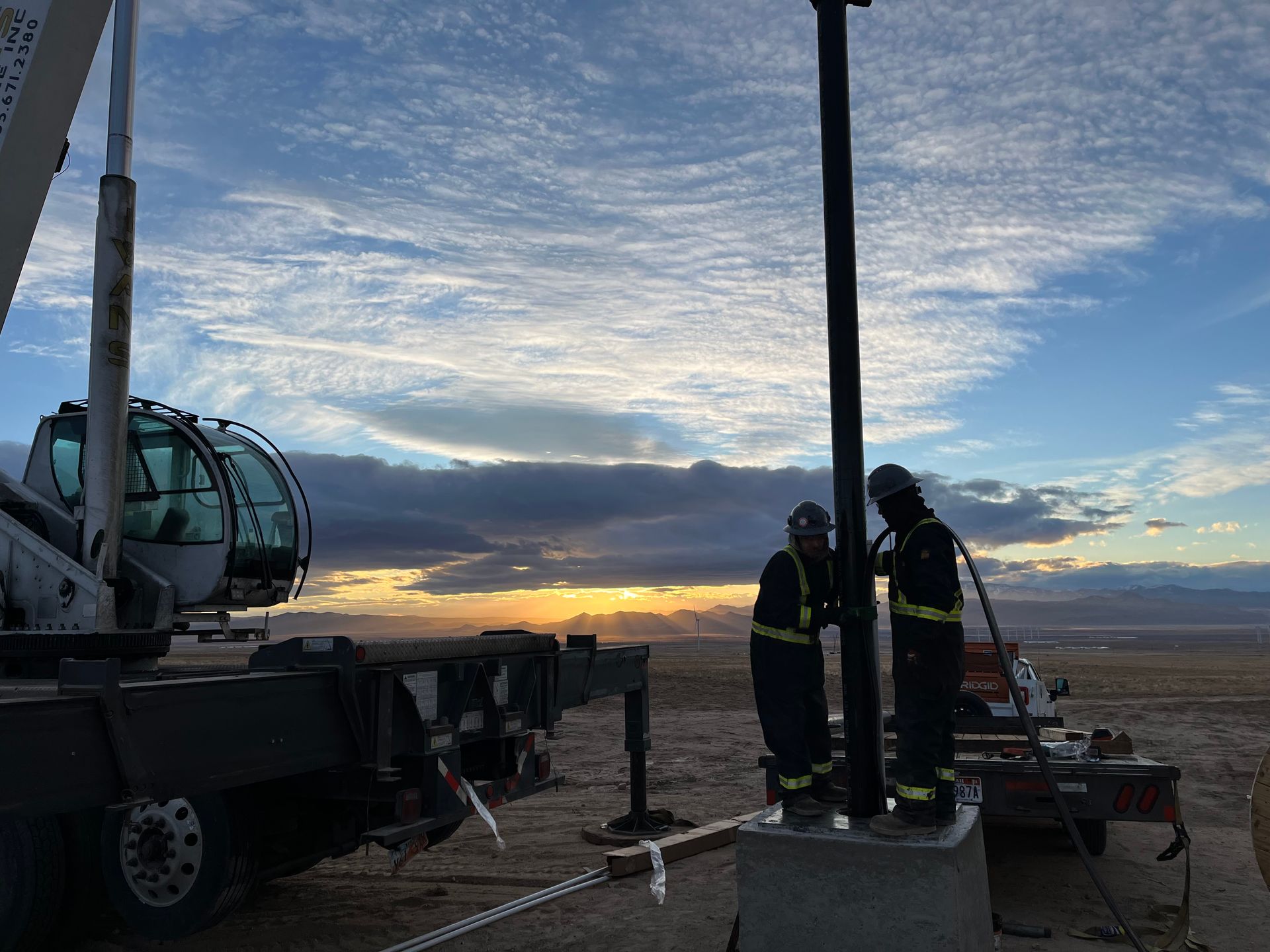 Two men are working on a pole in the desert near a crane.