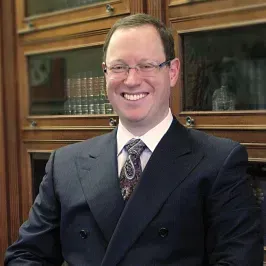 Man in a suit smiles, wearing glasses, in front of a bookshelf.