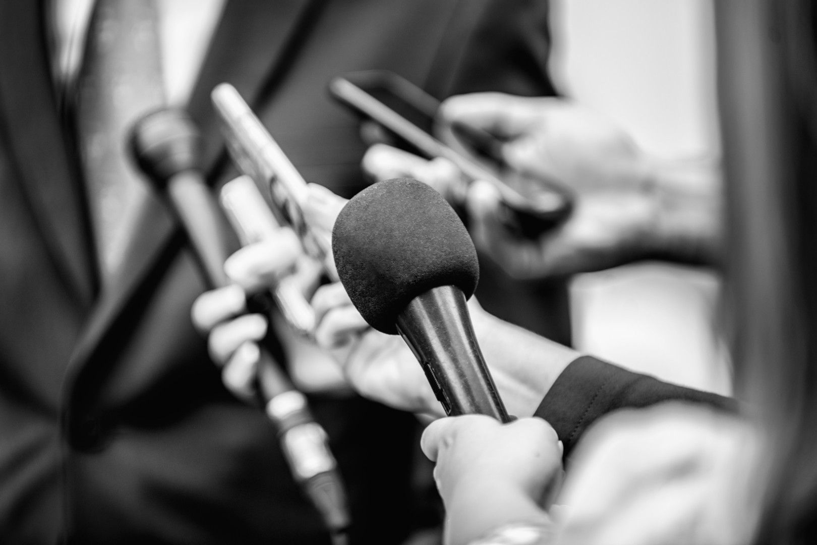 Torso of a man in a business suit with numerous microphones pointed at him.