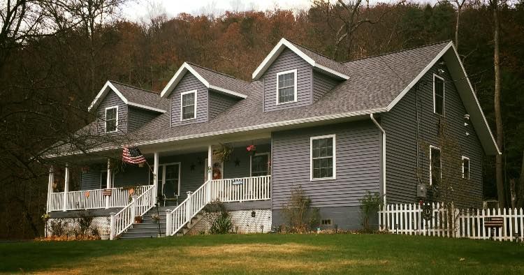 Gray house with white porch, picket fence, and dormer windows against a forested hillside.