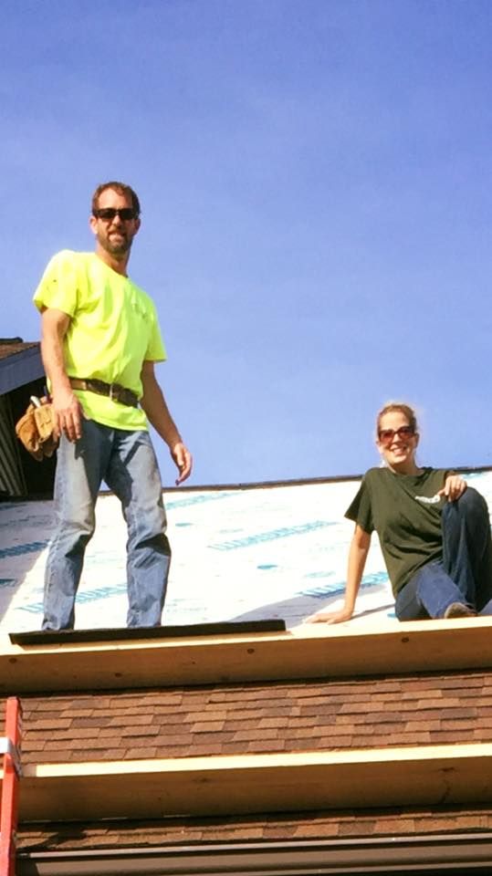 Two people on a roof with blue sky. Man in neon shirt and jeans, woman in green shirt, smiling.
